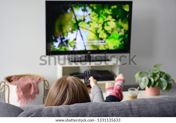 Young woman watching television with subtitles\
while sitting comfortably on sofa at home in living room. Nature,\
green, documentary, tv\
screen