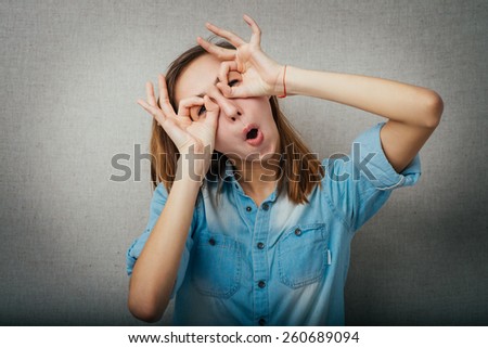 Young woman watching through fingers glasses over a grey background