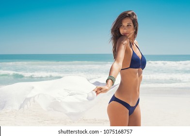 Young woman waving white scarf in wind at beach. Latin happy girl in blue bikini holding tissue and looking away at sea. Sexy fashion woman playing with the scarf on seashore. 