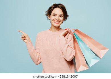 Young woman wear beige knitted sweater casual clothes hold in hand paper package bags after shopping point aside isolated on plain light pastel blue cyan background. Black Friday sale buy day concept
