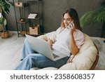 Young IT woman wear casual clothes sits in armchair hold use work on laptop pc computer talk on mobile cell phone stay home hotel flat rest spend free spare time in living room indoor. Lounge concept