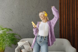 Young Woman Wear Casual Clothes Headphones Listen Music Dance Raise Up Hand Near Grey Sofa Couch Stay At Home Hotel Flat Rest Relax Spend Free Spare Time In Living Room Indoor People Lounge Concept