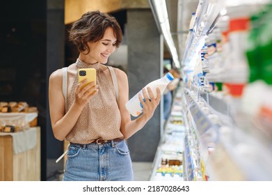 Young woman wear casual clothes backpack shopping at supermaket store buy choose dairy produce take milk read shelf life using mobile cell phone inside hypermarket Purchasing gastronomy food concept. - Shutterstock ID 2187028643