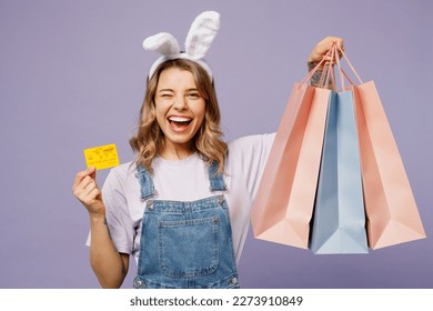 Young woman wear casual clothes bunny rabbit ears hold paper package bag after shopping credit bank card wink isolated on plain light pastel purple background studio Happy Easter sale buy day concept