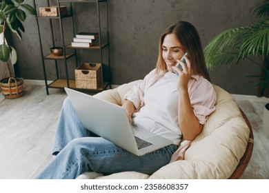 Young IT woman wear casual clothes sits in armchair hold use work on laptop pc computer talk on mobile cell phone stay home hotel flat rest spend free spare time in living room indoor. Lounge concept - Shutterstock ID 2358608357