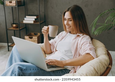 Young IT woman wear casual clothes sits in armchair hold use work on laptop pc computer drink tea stay home hotel flat rest relax spend free spare time in living room indoor. Lifestyle lounge concept - Powered by Shutterstock