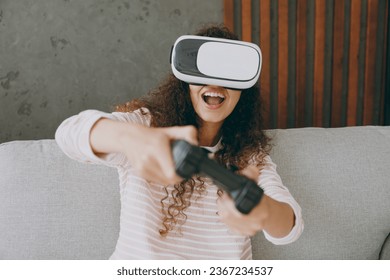Young woman wear casual clothes sits on grey sofa couch watch in vr headset pc gadget play pc game with joystick console stay at home hotel flat rest relax spend free spare time in living room indoor - Shutterstock ID 2367234537