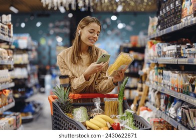 Young woman wear casual clothes earphones listen music use mobile cell phone hold pasta shopping at supermaket store grocery shop buy with cart choose products in hypermarket. Purchasing food concept - Powered by Shutterstock