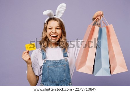 Young woman wear casual clothes bunny rabbit ears hold paper package bag after shopping credit bank card wink isolated on plain light pastel purple background studio Happy Easter sale buy day concept