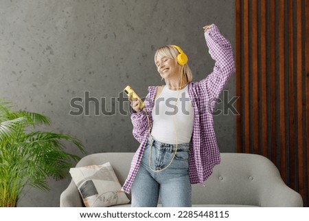 Young woman wear casual clothes headphones listen music dance raise up hand near grey sofa couch stay at home hotel flat rest relax spend free spare time in living room indoor People lounge concept
