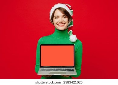 Young IT woman wear green turtleneck Santa hat posing hold use work on blank screen area laptop pc computer isolated on plain red background. Happy New Year 2024 celebration Christmas holiday concept