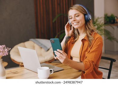 Young IT woman wear orange shirt headphones hold mobile cell phone use laptop pc computer listen music sit alone at table in coffee shop cafe indoor work or study. Freelance office business concept