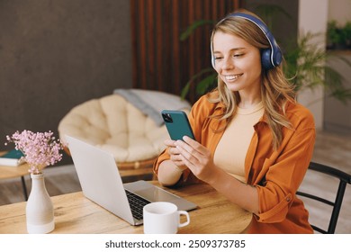 Young IT woman wear orange shirt headphones hold mobile cell phone using laptop pc computer listen music sit alone at table in coffee shop cafe indoor work or study. Freelance office business concept