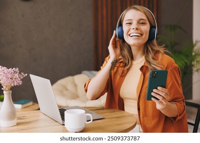 Young IT woman wear orange shirt headphones hold mobile cell phone use laptop pc computer listening music sit alone at table in coffee shop cafe indoor work or study. Freelance office business concept