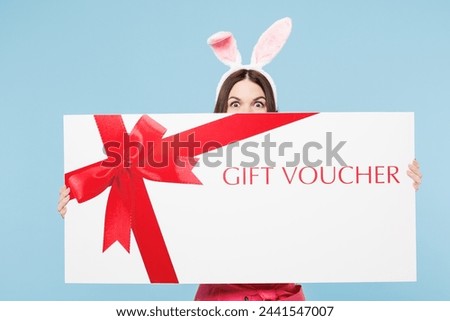 Young woman wear pink casual clothes rabbit bunny ears hold cover mouth with big huge gift coupon voucher card for store isolated on plain blue cyan background studio. Lifestyle Happy Easter concept