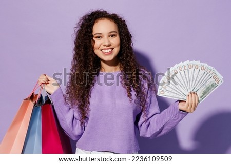 Young woman wear pullover hold in hand paper package bags after shopping fan of cash money in dollar banknotes isolated on plain pastel light purple color background Black Friday sale buy day concept