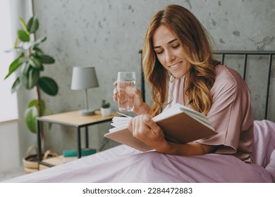 Young woman wear purple t-shirt pajama sit in bed read book drink water rest relax spend time in bedroom lounge home in hotel room wake up dream be lost in reverie good mood day. Real estate concept