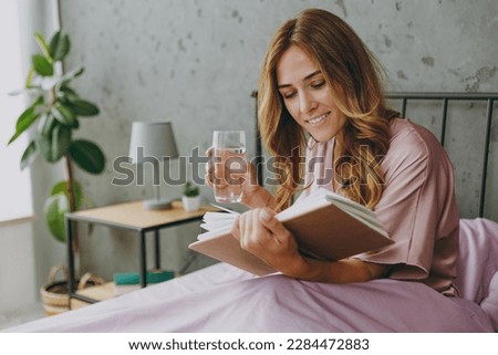 Young woman wear purple t-shirt pajama sit in bed read book drink water rest relax spend time in bedroom lounge home in hotel room wake up dream be lost in reverie good mood day. Real estate concept