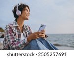 Young woman wear shirt casual clothes headphones sits listen to music use mobile cell phone listen music rest on sea ocean sand shore beach outdoor seaside in summer day free time. Lifestyle concept