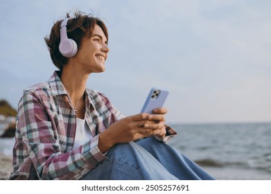 Young woman wear shirt casual clothes headphones sits listen to music use mobile cell phone listen music rest on sea ocean sand shore beach outdoor seaside in summer day free time. Lifestyle concept - Powered by Shutterstock