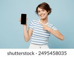 Young woman wear striped t-shirt casual clothes hold in hand use mobile cell phone point index finger on blank screen workspace area isolated on plain pastel light blue background. Lifestyle concept