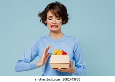 Young woman wear sweater hold a box with fastfood french fries potato burger show stop palm gesture reject say no isolated on plain pastel light blue background. People lifestyle junk food concept.