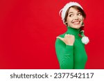 Young woman wear warm cozy green turtleneck Santa hat posing point thumb finger indicate on workspace area isolated on plain red background. Happy New Year 2024 celebration Christmas holiday concept