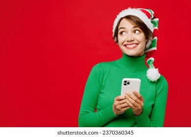 Young woman wear warm cozy green turtleneck Santa hat posing hold in hand use mobile cell phone look aside isolated on plain red background. Happy New Year 2024 celebration Christmas holiday concept - Powered by Shutterstock