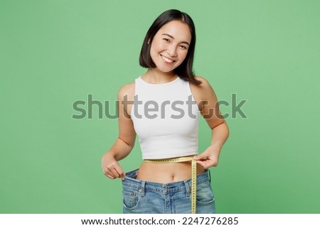 Young woman wear white clothes show loose pants after weightloss hold measure tape on waist isolated on plain pastel light green background. Proper nutrition healthy fast food unhealthy choice concept