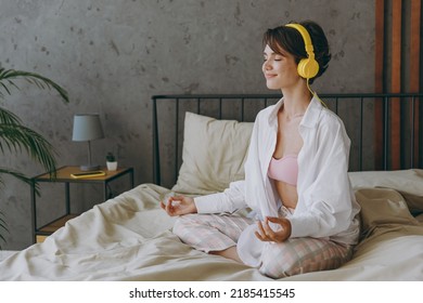 Young woman wear white shirt pajama headphones she lying in bed listen to music mantra meditating hold hands in yoga om gesture rest relax spend time in bedroom lounge home in own room hotel wake up - Powered by Shutterstock