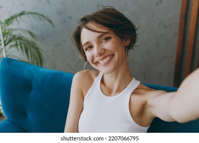 Young woman wear white tank shirt doing selfie shot pov on mobile cell phone sit on blue sofa couch stay at home hotel flat rest relax spend free spare time in living room indoor People lounge concept