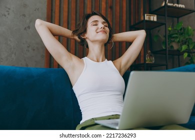 Young IT Woman Wear White Tank Shirt Hold Hands Behind Neck Use Work On Laptop Pc Computer Sit On Blue Sofa Stay Home Flat Rest Relax Spend Free Spare Time In Living Room Indoors People Lounge Concept