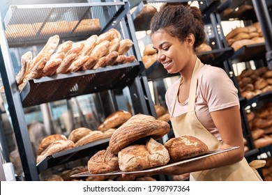Young woman wearing apron assistant at friendly bakery shop small business walking carrying tray of whole wheat bread loafs smiling cheerful - Powered by Shutterstock