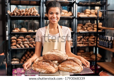Young woman wearing apron assistant at friendly bakery shop small business holding rye bread loafs posing to camera smiling happy