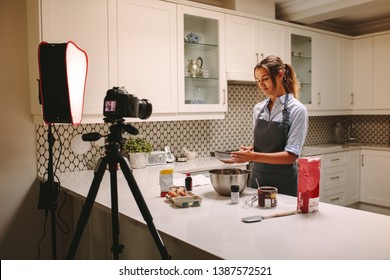 Young woman wearing apron filming herself preparing cake in the kitchen. Pastry chef recording content for the food and baking vlog using a camera mounted on tripod.