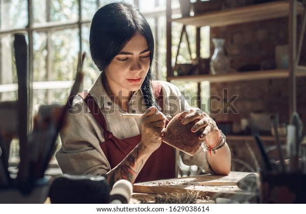 Young woman wearing
apron making pottery indoors at creative studio craftsperson
concept sitting at table using modeling tool creating details on
cup in the dark pensive