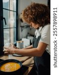 Young woman wearing apron pouring whisked eggs from bowl into frying pan while cooking omelette in domestic kitchen next to window