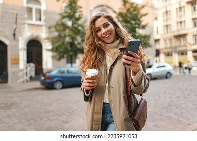 Young woman wearing autumn coat walking with smartphone and coffee cup in a city street - Powered by Shutterstock