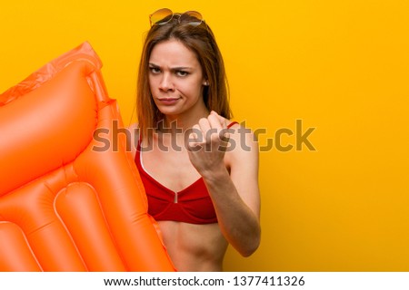Young woman wearing bikini, holding an air mattress bed showing fist to camera, aggressive facial expression.