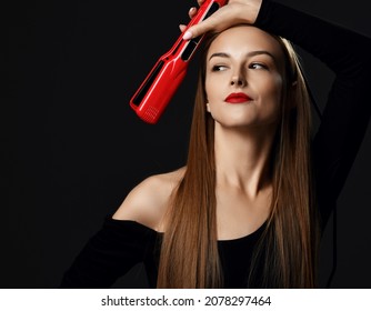 Young woman wearing black clothes skilled professional with silky hair holds hand with new red hair straightener at her head and looks aside over dark background. Haircare, beauty, wellness concept