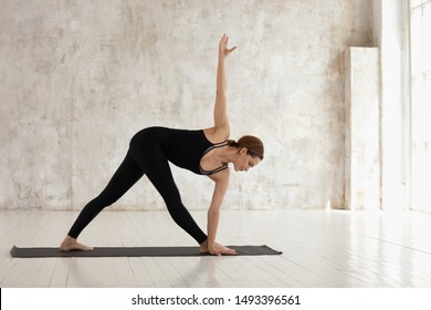 Young woman wearing black sportswear, practicing yoga, beautiful girl standing in Utthita Trikonasana pose on mat, extended triangle exercise, working out in modern yoga studio with wooden floor