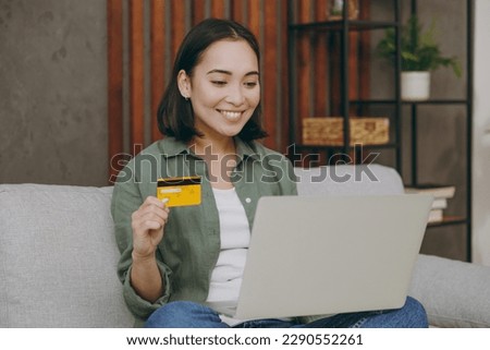 Young woman wearing casual clothes use laptop pc computer hold credit bank card shopping online sits on grey sofa couch stay at home hotel flat rest relax spend free spare time in living room indoor