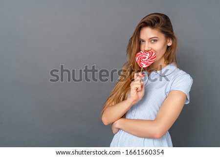 Young woman wearing casual dress studio isolated on gray wall covering lips with heart-shaped lollipop looking camera flirting