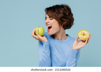 Young woman wearing casual sweater look camera hold donut dessert bite apple choose beatween healthy and junk food isolated on plain pastel light blue background studio. People lifestyle food concept