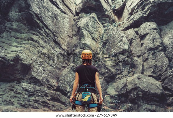 Young\
woman wearing in climbing equipment standing in front of a stone\
rock outdoor and preparing to climb, rear\
view