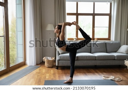 Young woman wearing comfy sportswear practicing asanas at home, standing in Natarajasana exercise or Lord of the Dance, working out alone at modern living room, full length view, yoga time concept
