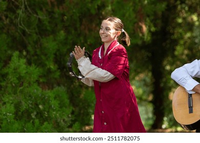 Young woman wearing a costume playing a tambourine and smiling in a park with a man playing guitar out of focus behind her - Powered by Shutterstock