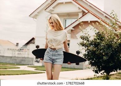 Young Woman Wearing Denim Shorts And White T Shirt, Holding A Long Board, Outdoors, On Street.