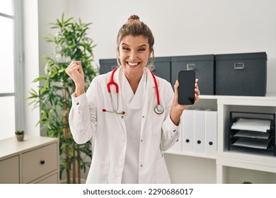 Young woman wearing doctor uniform holding smartphone screaming proud, celebrating victory and success very excited with raised arm 