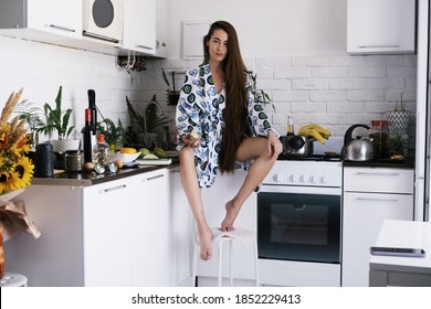 Young Woman Wearing Dressing Gown And White Underwear Sitting On The Desk, Eating Green Apple. Morning Kitchen Chaos